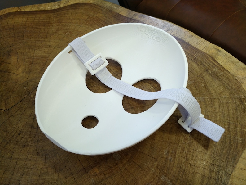 May include: White plastic face mask with two eye holes and a mouth hole. The mask has an adjustable strap with a buckle.