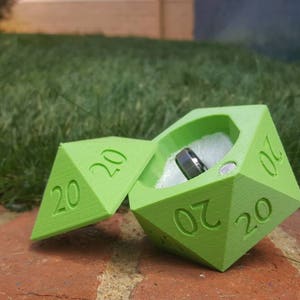 May include: A green 20-sided die ring box with a silver ring inside. The box is open and the ring is visible.