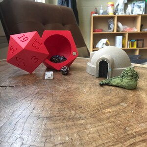 May include: A red dice tower with a dice tray, a white dice, and four multi-colored dice inside. A beige model of a dome-shaped building with a door and a green creature figurine are on a wooden table.