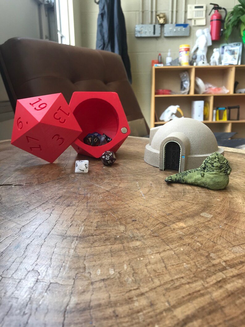 May include: A red dice tower with a dice tray, a white dice, and four multi-colored dice inside. A beige model of a dome-shaped building with a door and a green creature figurine are on a wooden table.