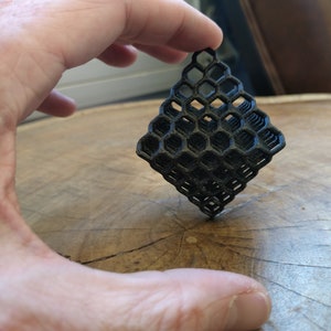 Puede incluir: Un objeto geom&eacute;trico de color negro impreso en 3D con un patr&oacute;n de panal de abeja. El objeto tiene forma de diamante y est&aacute; formado por varias capas.