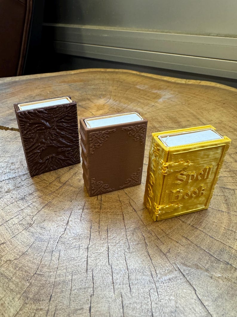 May include: Three small, decorative boxes with a book-like design. The boxes are brown, dark brown, and gold. The gold box has the words "Spell Book" printed on the front.