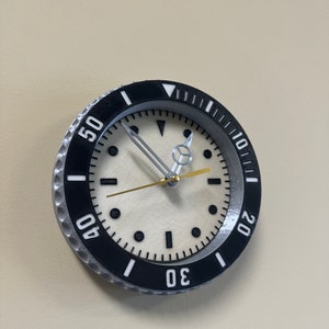 Puede incluir: Un reloj de pared negro y plateado con una esfera blanca y marcas negras. El reloj tiene un bisel grande con los n&uacute;meros 10, 20, 30, 40 y 50 alrededor del borde.