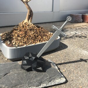 K&ouml;nnte beinhalten: Ein silbernes Schwert mit schwarzem Griff steht aufrecht auf einem schwarzen, geometrischen Stein. Das Schwert liegt auf einer grauen Schieferplatte. Im Hintergrund befindet sich ein Bonsai-Baum in einem grauen Topf.