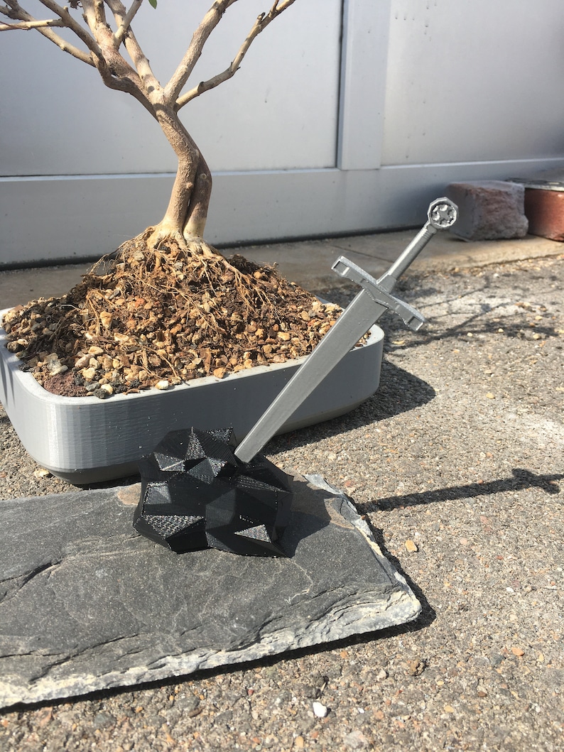 K&ouml;nnte beinhalten: Ein silbernes Schwert mit schwarzem Griff steht aufrecht auf einem schwarzen, geometrischen Stein. Das Schwert liegt auf einer grauen Schieferplatte. Im Hintergrund befindet sich ein Bonsai-Baum in einem grauen Topf.