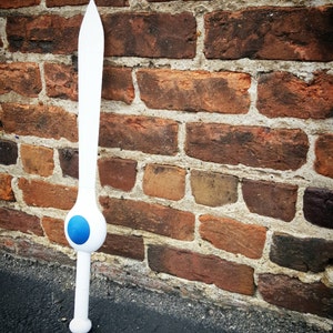 May include: A white wooden sword with a blue circle on the handle. The sword is leaning against a brick wall.