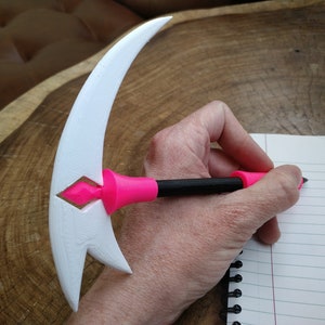 Puede incluir: Un bol&iacute;grafo blanco impreso en 3D con un mango rosa en forma de media luna. El bol&iacute;grafo se est&aacute; utilizando para escribir en un cuaderno de l&iacute;neas.