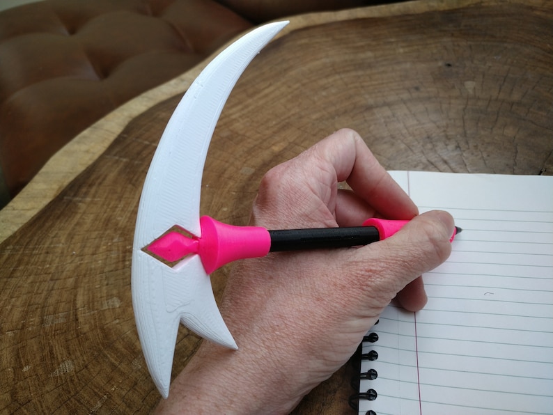 Puede incluir: Un bol&iacute;grafo blanco impreso en 3D con un mango rosa en forma de media luna. El bol&iacute;grafo se est&aacute; utilizando para escribir en un cuaderno de l&iacute;neas.