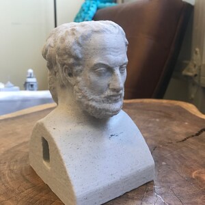 May include: A white plaster bust of a man with a beard. The bust is sitting on a wooden surface. The bust has a hole in the back.