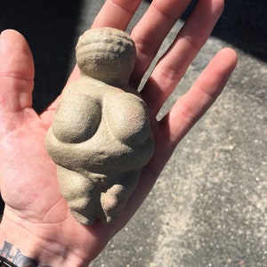 Puede incluir: Una peque&ntilde;a escultura de cer&aacute;mica beige que representa una figura femenina con rasgos exagerados. La escultura es lisa y tiene una superficie texturizada.