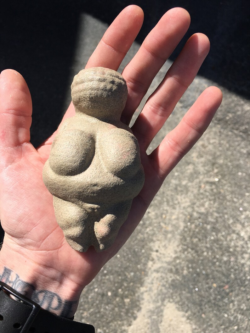 Puede incluir: Una peque&ntilde;a escultura de cer&aacute;mica beige que representa una figura femenina con rasgos exagerados. La escultura es lisa y tiene una superficie texturizada.