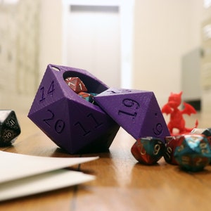 May include: A purple 20-sided die with a hinged lid, open to reveal smaller dice inside. The die is on a wooden surface with other dice, including a red dragon figurine.