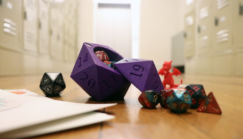 May include: A purple 20-sided die with a hinged lid, open to reveal smaller dice inside. The die is on a wooden surface with other dice, including a red dragon figurine.