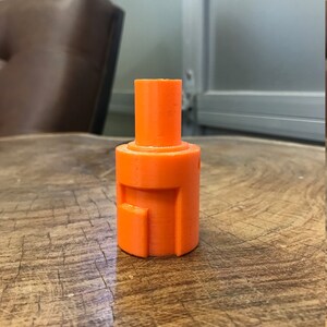 May include: An orange plastic 3D printed object with a cylindrical shape and a square opening on the side.