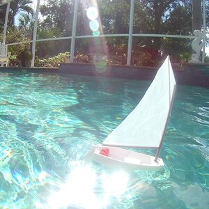 Puede incluir: Un velero de juguete blanco flota en una piscina azul. El velero tiene una vela blanca y una franja roja en el casco.