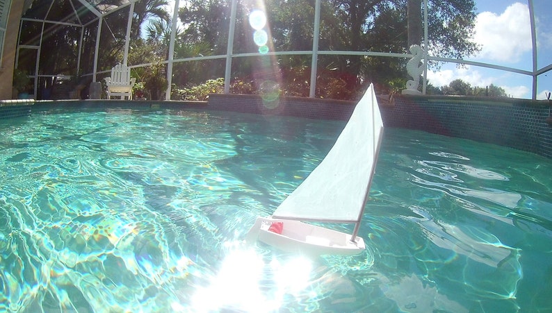Puede incluir: Un velero de juguete blanco flota en una piscina azul. El velero tiene una vela blanca y una franja roja en el casco.