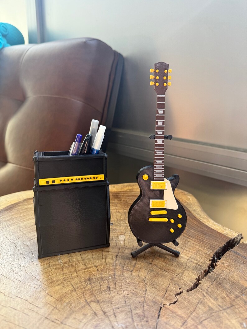 May include: A black and yellow electric guitar model on a stand next to a black amplifier with a pen holder. The amplifier has a yellow stripe and a row of knobs.