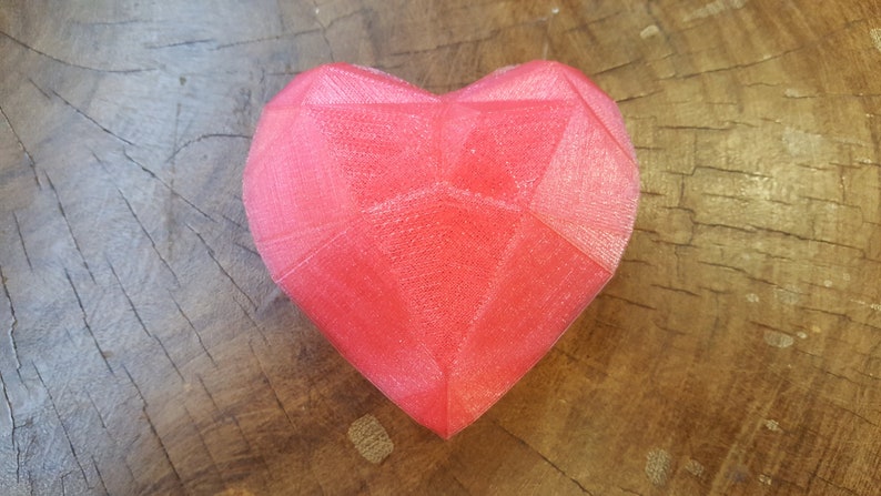 Puede incluir: Un objeto en forma de coraz&oacute;n rojo facetado. El coraz&oacute;n tiene un dise&ntilde;o geom&eacute;trico con m&uacute;ltiples superficies planas. El objeto descansa sobre una superficie de madera marr&oacute;n texturizada, creando un contraste de color y textura.