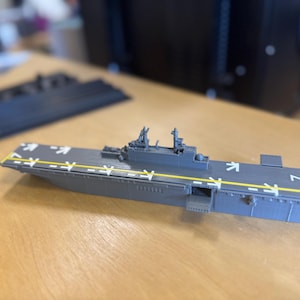 May include: A grey model aircraft carrier with white markings and yellow stripes. The miniature warship has detailed structures and is set on a wooden surface. The model is a representation of a naval vessel.