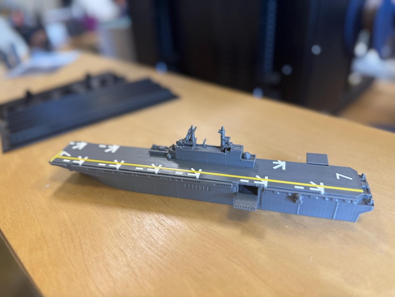 May include: A grey model aircraft carrier with white markings and yellow stripes. The miniature warship has detailed structures and is set on a wooden surface. The model is a representation of a naval vessel.