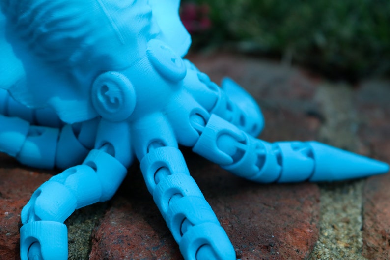 May include: A blue articulated octopus toy with segmented arms and a pointed tip on the end of one arm. The octopus is lying on a brick surface.