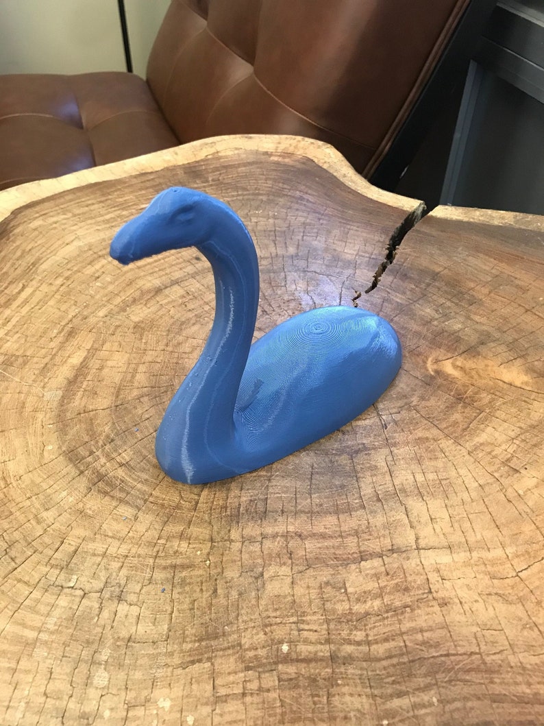 K&ouml;nnte beinhalten: Eine blaue 3D-gedruckte Skulptur einer mythischen Kreatur, die einem langhalsigen Dinosaurier &auml;hnelt, auf einer Holzoberfl&auml;che.
