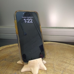 Puede incluir: Un smartphone con una funda transparente se muestra en un soporte de tel&eacute;fono de madera natural. La pantalla del tel&eacute;fono muestra la fecha y la hora. El soporte est&aacute; tallado en madera clara y tiene una forma org&aacute;nica &uacute;nica.