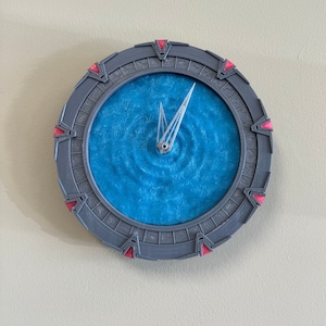 May include: A grey and red clock shaped like the Stargate from the TV show Stargate SG-1. The clock face is a blue swirling pattern.