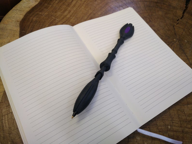 K&ouml;nnte beinhalten: Ein schwarzer Stift mit einer violetten Spitze und einem einzigartigen, strukturierten Design. Der Stift liegt offen auf einem karierten Notizbuch.