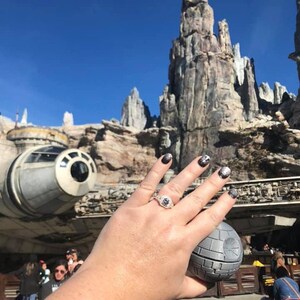 May include: A close-up of a hand wearing a silver ring with a large diamond, holding a small, round, gray object. The hand is in front of a large, gray, rocky structure and a white spaceship.