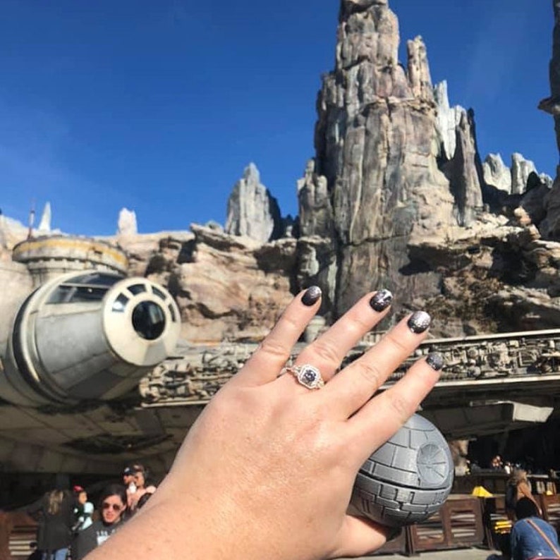May include: A close-up of a hand wearing a silver ring with a large diamond, holding a small, round, gray object. The hand is in front of a large, gray, rocky structure and a white spaceship.