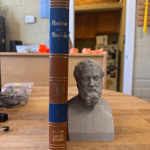 May include: A brown and blue hardcover book with the title "Herodotus - Thucydides" and the text "Great Books" on the spine. The book is next to a gray bust of a man with a beard.