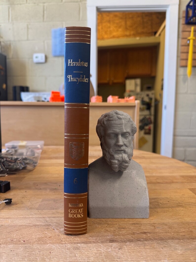 May include: A brown and blue hardcover book with the title "Herodotus - Thucydides" and the text "Great Books" on the spine. The book is next to a gray bust of a man with a beard.