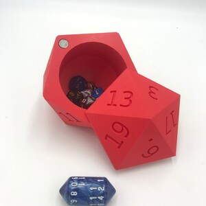 May include: A red, 20-sided dice box with a hinged lid. The box is open and has several dice inside. A blue, 10-sided dice is sitting on a white surface.