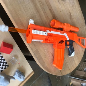 May include: An orange and white Nerf toy gun with a black grip and a scope. The gun has a magazine and a barrel with a white tip.