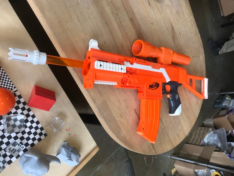 May include: An orange and white Nerf toy gun with a black grip and a scope. The gun has a magazine and a barrel with a white tip.