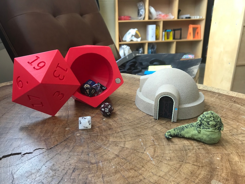 May include: A red, 3D printed dice tower with a dice tray, a white dice, a brown and black dice, and a beige 3D printed model of a dome-shaped building with a door. A green, 3D printed model of a creature is in the foreground.
