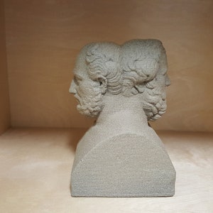 May include: A two-headed bust sculpture made of a light gray material. The sculpture is in a wooden shelf.