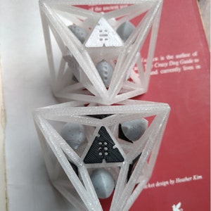 May include: Two stacked, translucent white 3D-printed geometric octahedron cages. Each cage contains smaller, loose, grey and white triangular and spherical components. The top cage features a white triangular insert, while the bottom cage has a black one.