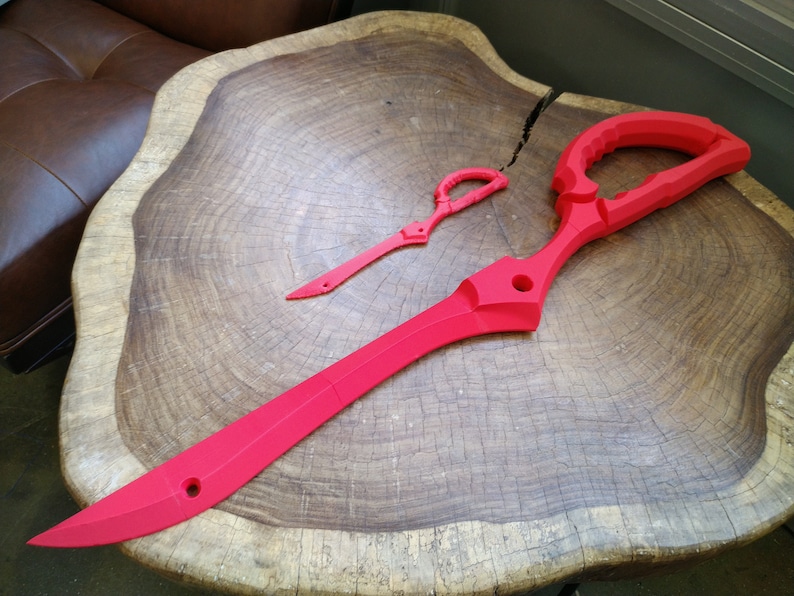 May include: Two red plastic prop swords, one large and one small, are displayed on a wooden surface. The larger sword is shaped like a pair of scissors.