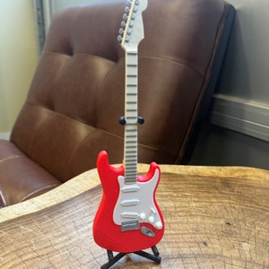May include: A red electric guitar with a white pickguard and a black stand. The guitar has a white neck and silver tuning pegs.