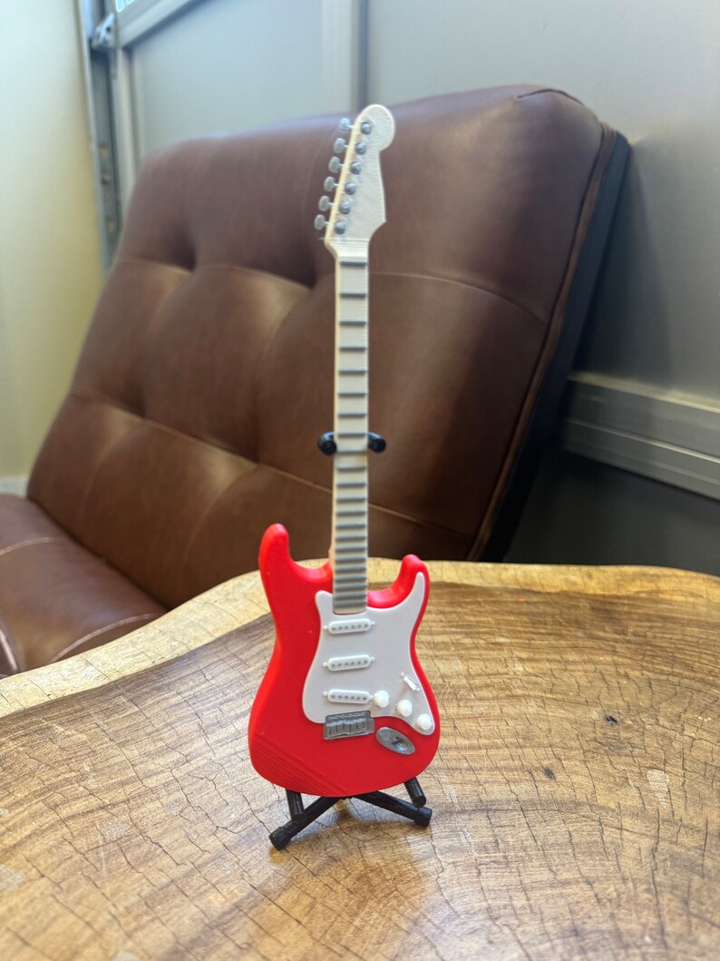 May include: A red electric guitar with a white pickguard and a black stand. The guitar has a white neck and silver tuning pegs.