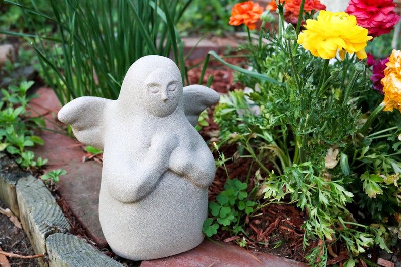 K&ouml;nnte beinhalten: Eine wei&szlig;e Steinengelstatue mit Fl&uuml;geln, die in einem Garten mit gr&uuml;nen Pflanzen und rotem Mulch steht. Der Engel betet mit ineinander verschr&auml;nkten H&auml;nden.