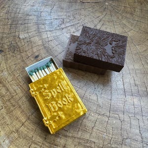 May include: A gold matchbox with the words "Spell Book" on the front. The matchbox is sitting on a wooden surface next to a brown matchbox with a textured design.