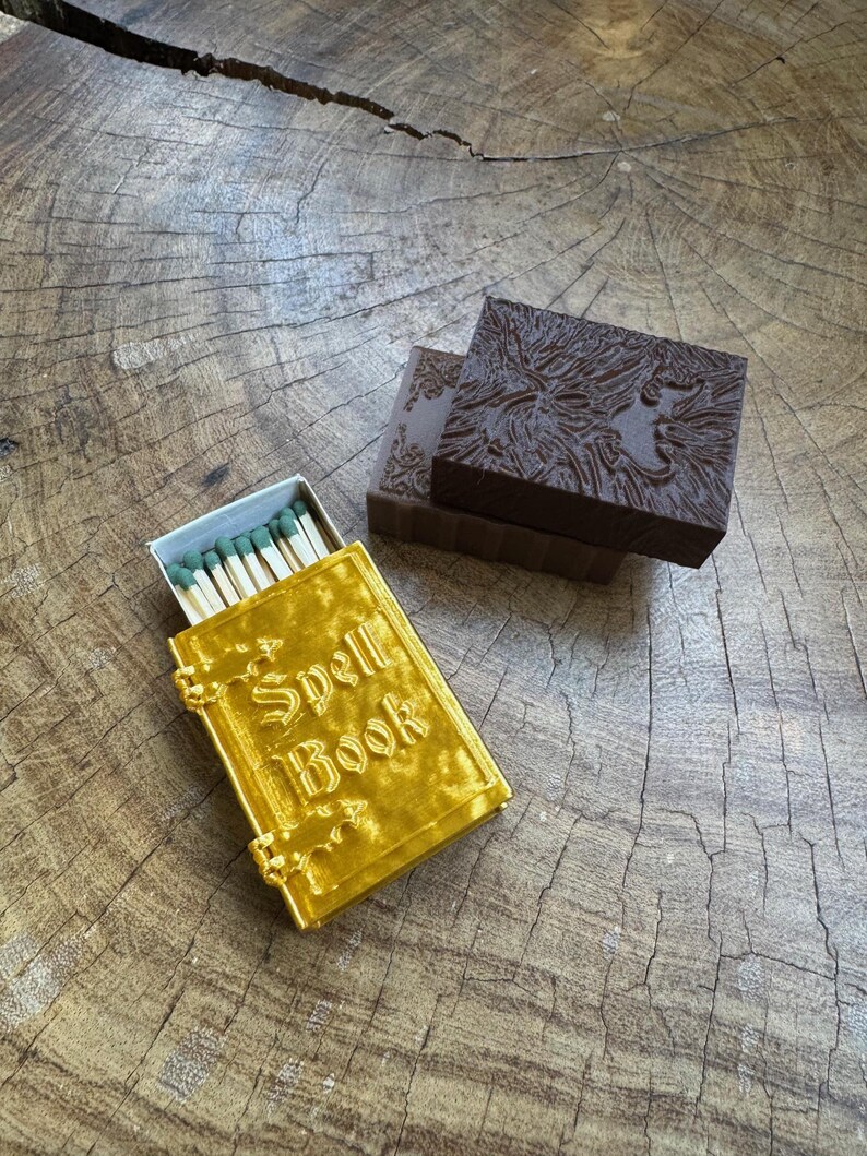 May include: A gold matchbox with the words "Spell Book" on the front. The matchbox is sitting on a wooden surface next to a brown matchbox with a textured design.