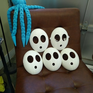 May include: Five white plastic masks with black eye holes and a single mouth hole, arranged in a pyramid shape on a brown leather chair. A blue octopus toy is in the background.