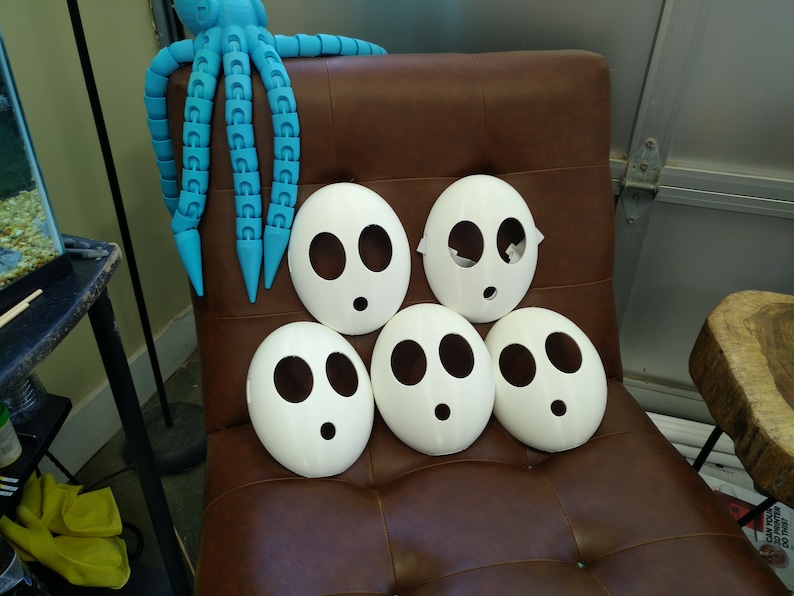 May include: Five white plastic masks with black eye holes and a single mouth hole, arranged in a pyramid shape on a brown leather chair. A blue octopus toy is in the background.