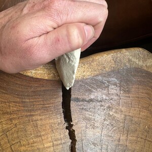 May include: A hand holding a light gray stone tool over a wooden surface with a crack running down the center.