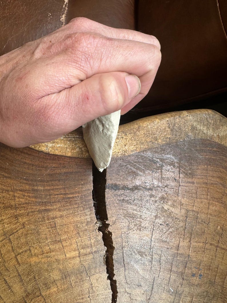 May include: A hand holding a light gray stone tool over a wooden surface with a crack running down the center.