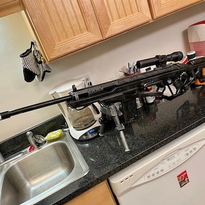 May include: A black sniper rifle with a scope mounted on top, resting on a tripod in a kitchen setting. The rifle is positioned on a black countertop with a stainless steel sink and a white coffee maker in the background.